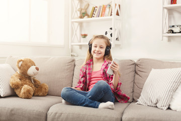 Happy little female child and her teddy bear listening to music on sofa at home