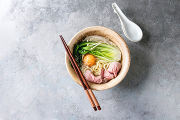 Asian dish udon noodles with egg yolk, sesame, mushrooms, boc choy, sliced sous vide cooked meat served in ceramic bowl with spoon and chopsticks over grey texture background. Top view, space.