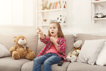 Scared casual little girl watching tv while sitting on sofa at home