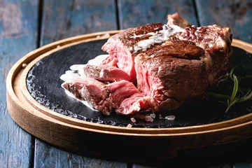 Papier Peint photo autocollant Steakhouse Sliced grilled sous-vide black angus beef tomahawk steak on bone served with salt, pepper, rosemary and white sauce on round wooden slate cutting board over dark wooden plank background. Close up