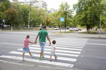 In the summer on the street at the pedestrian crossing father and son and daughter cross the road. Dad keeps the children by the hands.