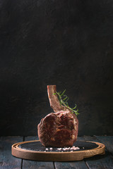 Grilled black angus beef tomahawk steak on bone served with salt, pepper and rosemary on round slate cutting board over dark wooden plank kitchen table. Copy space.