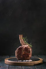 Poster Steakhouse Grilled black angus beef tomahawk steak on bone served with salt, pepper and rosemary on round slate cutting board over dark wooden plank kitchen table. Copy space.