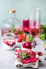 Vegan fruit salad with berries and mint served in pink dragon fruit on cloth with jars of yogurt, bottle of lemonade, ingredients above on grey table. Healthy eating