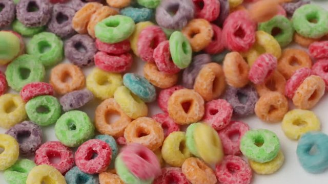 Very close video of pouring generic dry sugar coated fruity flavored breakfast cereal into a large white bowl.