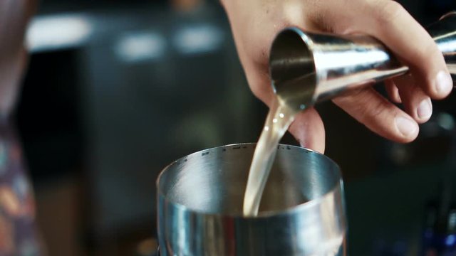 Close up of yellow liquer is being poured to steel shaker from measure glass in modern bar, slow motion. Barmen is inclining the cup by his hand and delicious drink is falling to the equipment for
