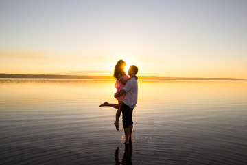 Fototapeta premium Young couple is embracing in the water on summer beach. Sunset over the sea.Two silhouettes against the sun. Just married couple hugging. Romantic love story. Man and woman in holiday honeymoon trip.