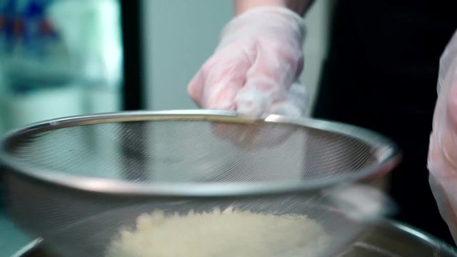 Wheaten flour is being bolted through sieve before cake cooking, close up. Big stainer is being shaken by confectioner in white latex gloves and powder is falling down to the steel bowl, where dough