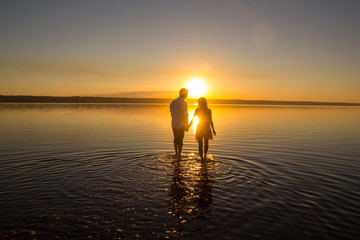 Fototapeta premium Young couple is walking in the water on summer beach. Sunset over the sea.Two silhouettes against the sun. Just married couple hugging. Romantic love story. Man and woman in holiday honeymoon trip.