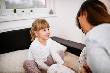 Adorable blonde little girl having fun with mother. Sitting in pajamas on the bed.