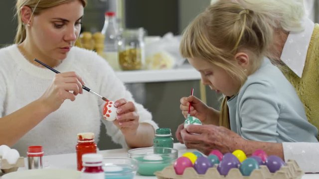 Little girl sitting on knees of grandmother, painting eggs for Easter dinner and talking with mother