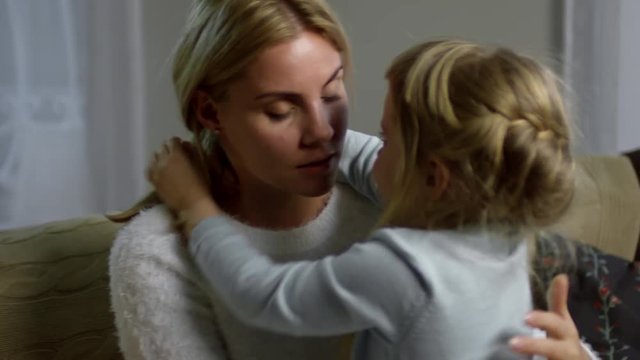 Beautiful mother talking to little daughter, stroking her hair and hugging with love while sitting on sofa at home