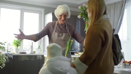 Joyous grandmother greeting her daughter and little granddaughter in the kitchen: woman and girl hugging senior lady and giving her flowers for Mothers Day - Powered by Adobe