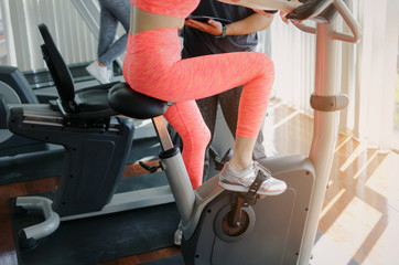 midsection of young woman slim body exercising on bicycle machine with young man personal trainer in fitness gym at morning, bodybuilder, healthy lifestyle, fitness, workout and sport training concept