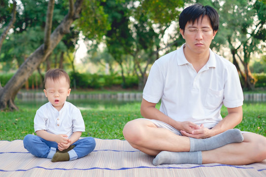 Father with eyes closed and Cute little Asian 18 months / 1 year old toddler baby boy child practices yoga & meditating outdoors on nature in summer, Healthy lifestyle concept, Selective focus at kid
