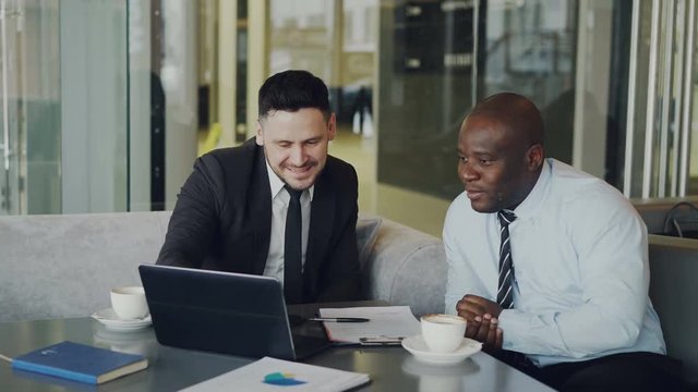 Two multi ethnic businessmen looking at laptop computer and laughing in glassy cafe. Business collaegues having fun and joking