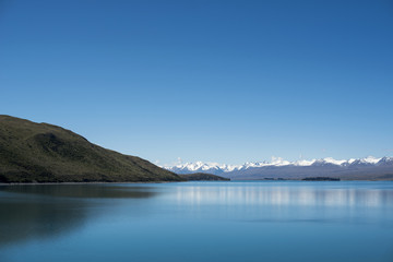 Paisaje de picos de montañas nevados con cielo azul y nubes frente un lago y valle con árbol en el Monte Cook, Nueva Zelanda.