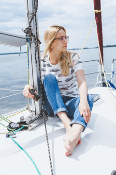 slender barefooted blonde in spectacles sits on nose of yacht