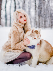 Pleasing girl in a fur coat sitting with her dog in a winter forest