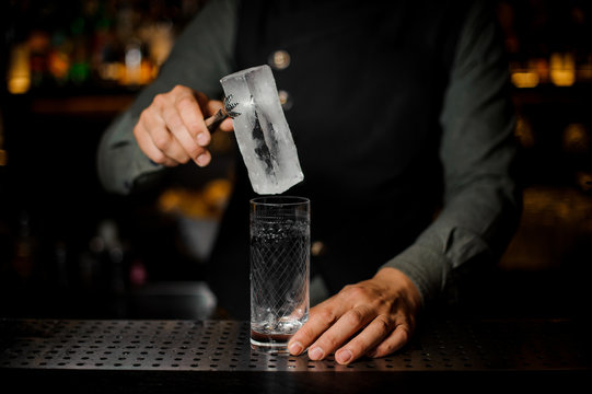 Bartender putting a big ice cube into an empty glass on a bar counter