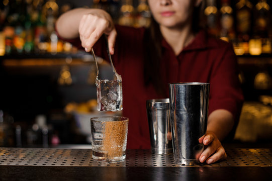 Barmen girl putting a big ice cube into an empty glass on a bar counter