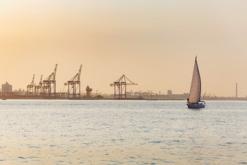 Beautiful seascape. Lonely boat with sails in a calm sea with reflection of sunlight near a industrial seaport on the coastline against the background of a clear warm evening sunset