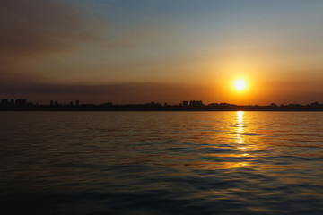 Beautiful sea landscape. Bright warm sunset in the sea near the coastline with the city's silhouettes. Sun hides behind the house. Colors of the sky are reflected in the water