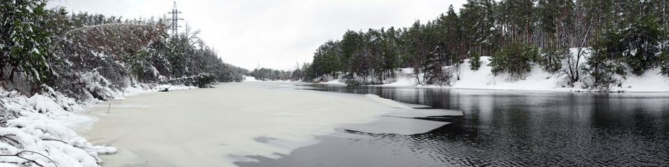 Panorama Beautiful natural landscape. Half frozen and ice-covered winter wide river in the forest with coniferous trees, bushes and snow drifts on the both banks