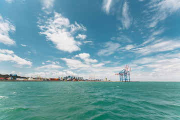 Beautiful seascape. Azure wavy restless sea with splashes and foam on background a cloudy blue sky on the horizon industrial large seaport on the shoreline