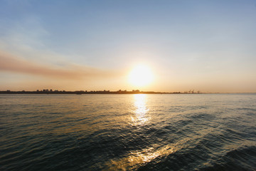 Beautiful sea landscape. Bright warm sunset in the sea near the coastline with the city's silhouettes. Sun hides behind the house. Colors of the sky are reflected in the water