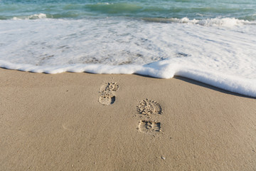 Footprints of a man are stamped on a sandy beach. Surf, waves with foam and spray wash off the tracks. World of nature, environmental