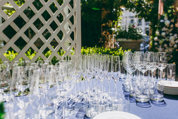 Preparing for party. Many clean shiny empty glasses for wine, champagne, whiskey, juice, water and other drinks on the table with a blue tablecloth in the gazebo at an outdoor party in the garden