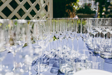 Preparing for party. Many clean shiny empty glasses for wine, champagne, whiskey, juice, water and other drinks on the table with a blue tablecloth in the gazebo at an outdoor party in the garden