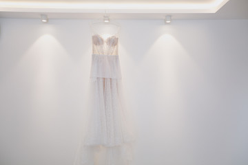 A beautiful tender elegant wedding dress with lace and thin straps for the bride hangs on a hanger against the background of a white wall and light bulbs. Wedding wear, accessories