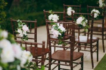 Beautiful decoration of a wedding ceremony in a green autumn garden. Brown vintage wooden chairs for guests decorated with boutonniere of white and pink peonies and green leaves
