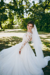 Fototapeta na wymiar Beautiful wedding photosession. The smiling bride in a white lace dress with a plume, long sleeves, hairpin in hair, white veil turns around in a large green garden on weathery sunny day