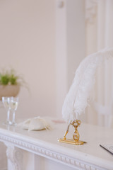 Elements of wedding decor Wide white feather for painting on a gold stand with decoration in the form of a flower on a shelter near glasses and pillow for rings in the white interior