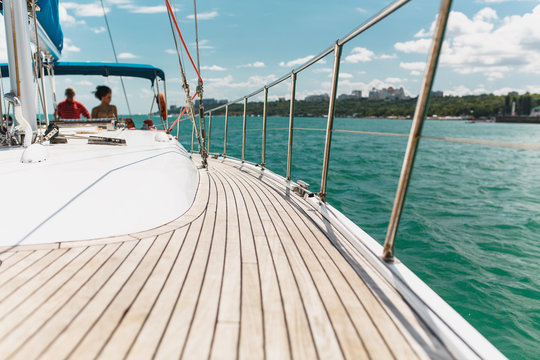 Sailing boat deck on a white yacht with teak wooden deck, metal railing, set of red ropes with people on board in the sea near the coastline in the background blue sky with clouds in sunny day