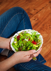 Green vegan breakfast meal in bowl with various fresh mix salad leaves and tomato. Girl in jeans holding fork with knees and hands visible, top view on wooden background