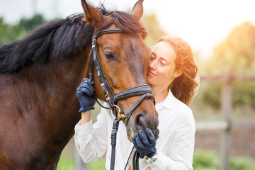 Young happy rider woman with her bay horse. Portrait of smiling girl with her stallion in paddock on sunny day