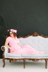 Young beautiful pregnant girl in a pink dress on a black background sitting on a vintage sofa