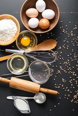 Fototapeta na wymiar Baking ingredients. Bowl, eggs, flour, eggbeater, rolling pin and eggshells on black chalkboard from above.