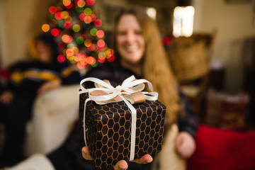 Woman holds beautifully wrapped present with Christmas tree lights in the background