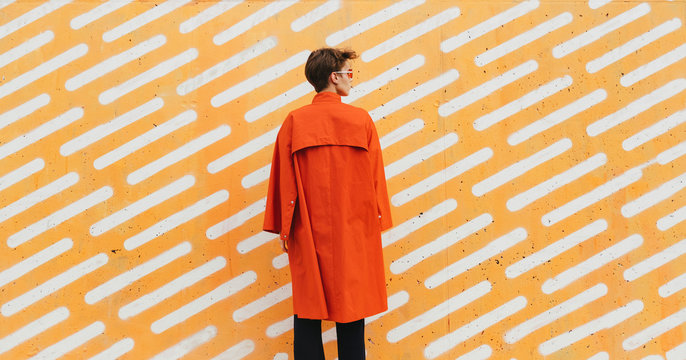 Urban stylish short hair female model in red/orange coat in front of orange background.