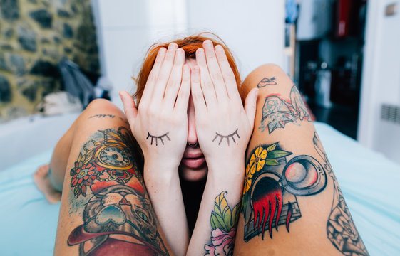 woman covering her face with drawn eyelashes on her hands between another womans tattoed legs