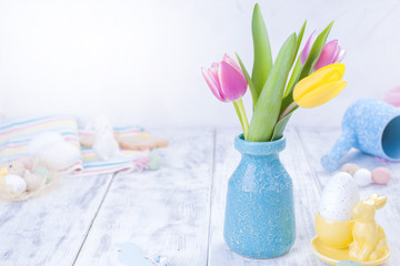 Spring tulips are pink and yellow. Different colors of eggs and pottery rabbit. Decor for Easter. White wooden background and place for text