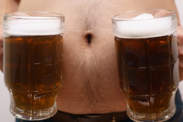 Beer belly, overweight. Man with obese abdomen holding two mugs of beer, close up. Alcoholism, unhealthy lifestyle