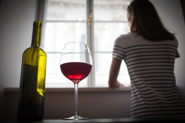 Woman looking out her window in a dark room drinking glass of wine. Sadness depression, alcoholism, loneliness concept. 