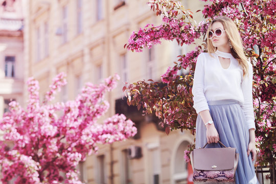 Outdoor portrait of young beautiful girl posing in street of european city, blooming trees on background. Model wearing stylish sunglasses, holding pink bag. Female fashion concept. Copy, empty space