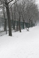 View of a wooden bench and a walk way by a green fence and row of trees covered in snow  during winter season, vintage toned
